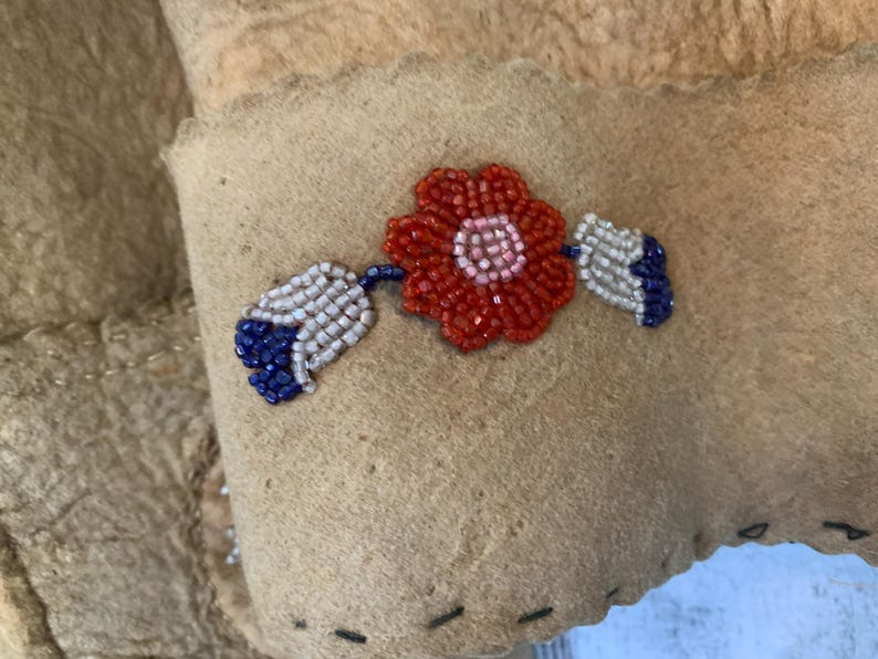 Puede incluir: Primer plano de un art&iacute;culo de ante color canela con un dise&ntilde;o floral de cuentas. La flor es roja con un centro rosa, y las hojas son blancas y azules. El art&iacute;culo tiene un borde cosido.