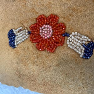 Puede incluir: Primer plano de una tela beige con un dise&ntilde;o floral de cuentas. La flor es roja con un centro rosa, y las hojas son blancas y azules. Las cuentas son peque&ntilde;as y crean un efecto texturizado.