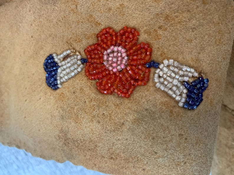 Puede incluir: Primer plano de una tela beige con un dise&ntilde;o floral de cuentas. La flor es roja con un centro rosa, y las hojas son blancas y azules. Las cuentas son peque&ntilde;as y crean un efecto texturizado.