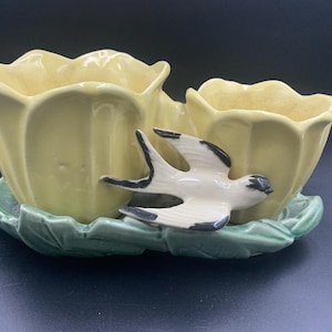 May include: A ceramic planter featuring two yellow tulip-shaped vessels attached to a green leaf-shaped base. A small, cream-coloured bird with black wing and head markings is attached to the front of the vessels, appearing to fly across the surface.