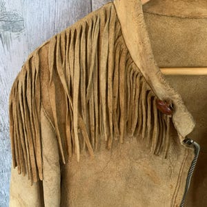 Puede incluir: Chaqueta de ante color canela con flecos en los hombros. La chaqueta tiene cierre de cremallera y un peque&ntilde;o bot&oacute;n redondo en el cuello. La chaqueta est&aacute; colgada en una percha de madera.