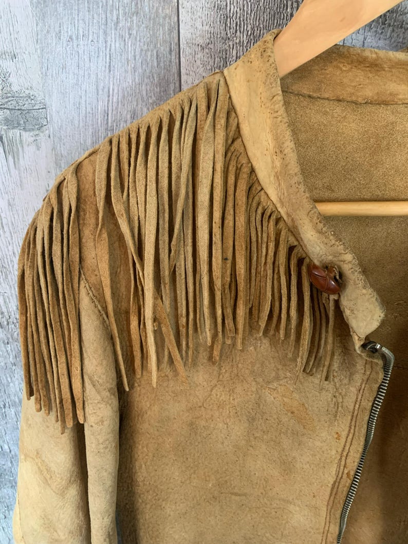 Puede incluir: Chaqueta de ante color canela con flecos en los hombros. La chaqueta tiene cierre de cremallera y un peque&ntilde;o bot&oacute;n redondo en el cuello. La chaqueta est&aacute; colgada en una percha de madera.