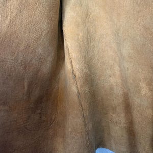 Puede incluir: Primer plano de un trozo de cuero de color marr&oacute;n claro. El cuero tiene una superficie texturizada con grano visible y una costura cosida en el centro. Los bordes del cuero son irregulares.