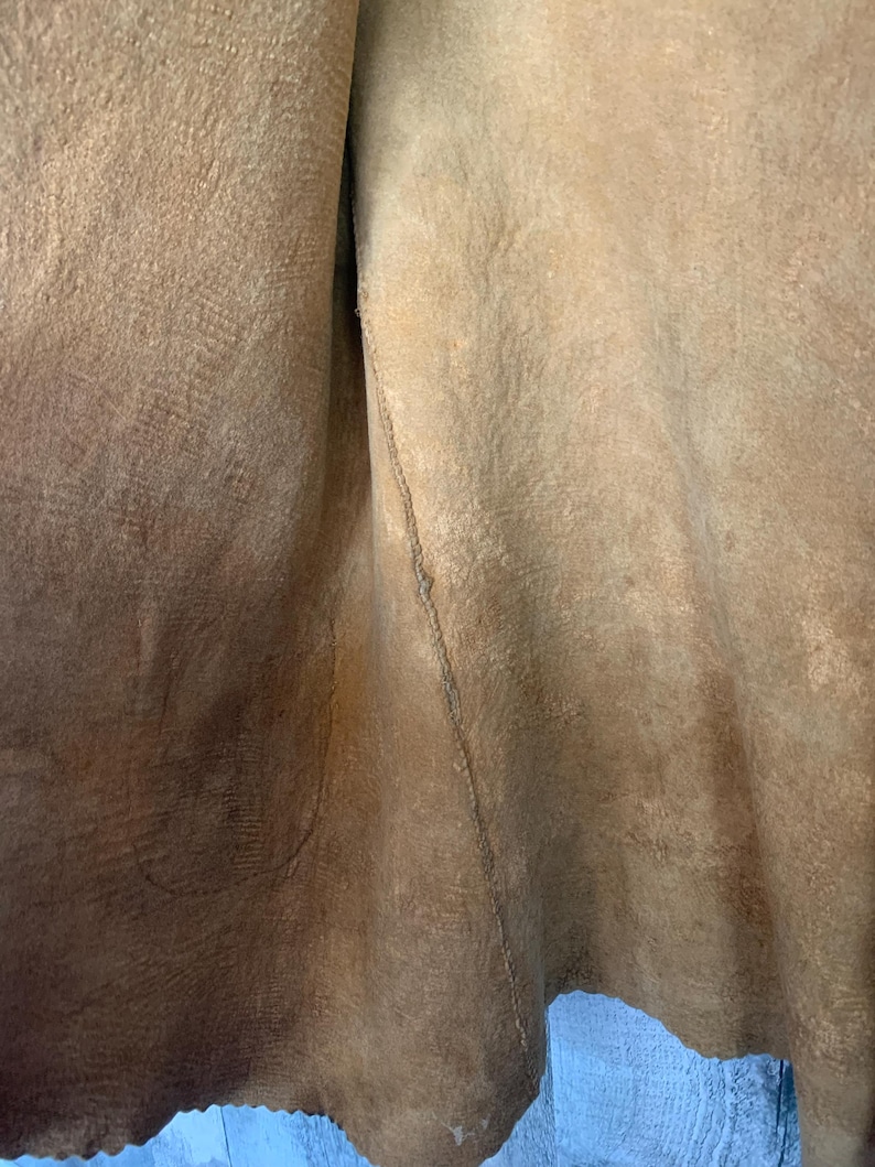 Puede incluir: Primer plano de un trozo de cuero de color marr&oacute;n claro. El cuero tiene una superficie texturizada con grano visible y una costura cosida en el centro. Los bordes del cuero son irregulares.