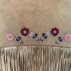 Puede incluir: Primer plano de una prenda de ante color canela con bordado floral de cuentas. Las flores son en tonos rosa, rojo y naranja, con detalles azules y blancos. El borde inferior presenta flecos.