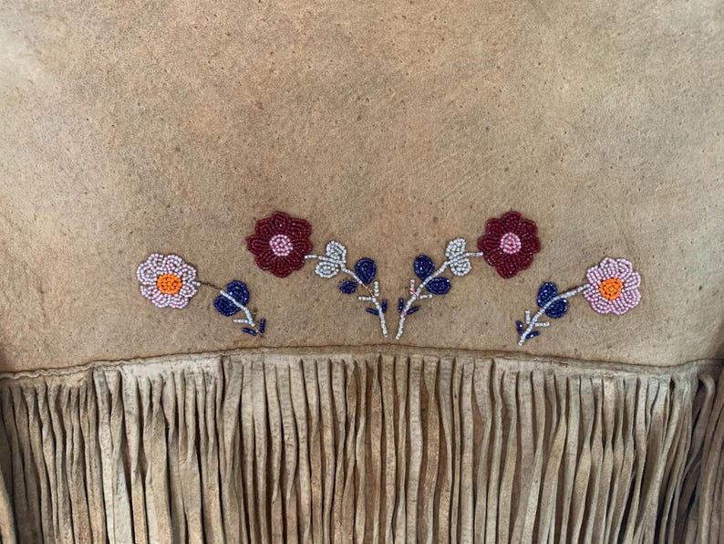 Puede incluir: Primer plano de una prenda de ante color canela con bordado floral de cuentas. Las flores son en tonos rosa, rojo y naranja, con detalles azules y blancos. El borde inferior presenta flecos.