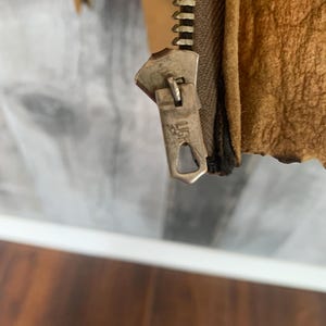 Puede incluir: Primer plano de la cremallera de una chaqueta de cuero marr&oacute;n. La cremallera es de color plateado y tiene la palabra "LIG" grabada en el tirador. El cuero de la chaqueta tiene un aspecto texturizado.