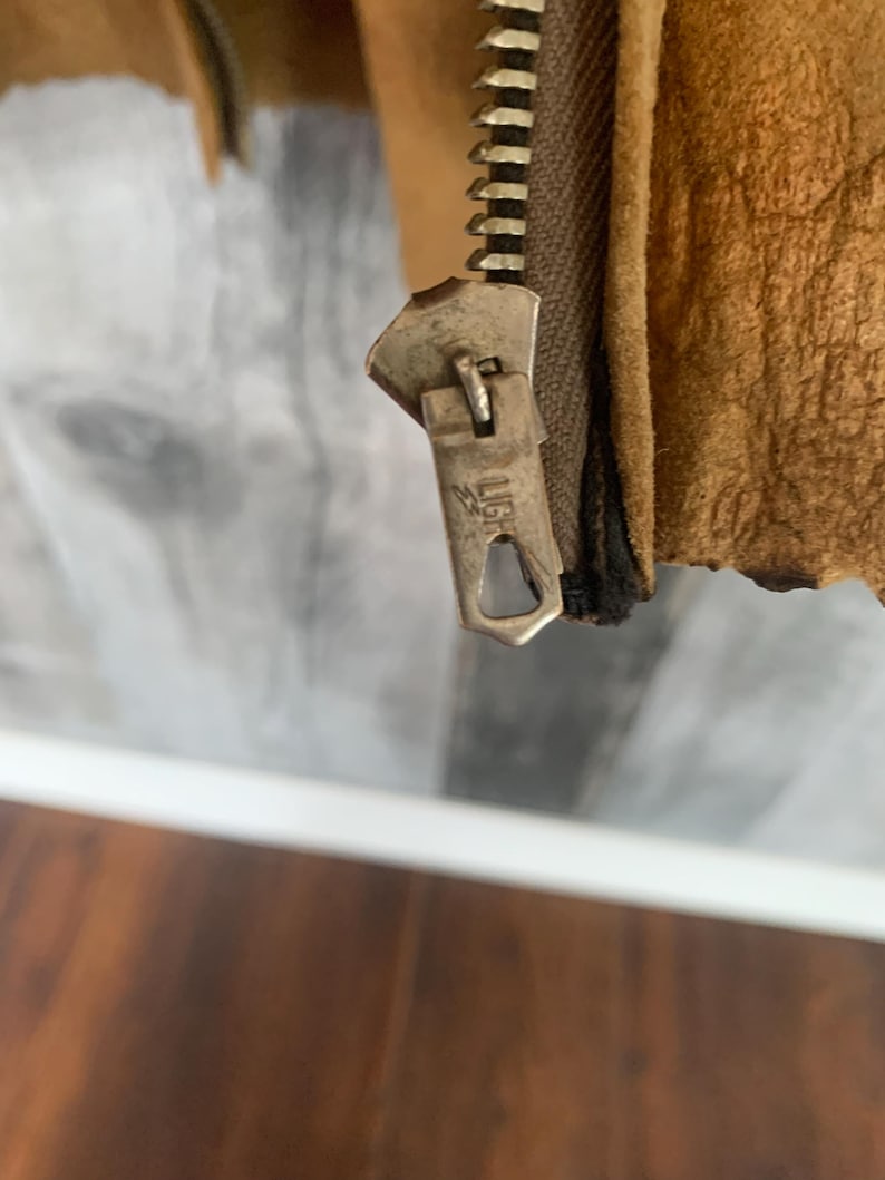 Puede incluir: Primer plano de la cremallera de una chaqueta de cuero marr&oacute;n. La cremallera es de color plateado y tiene la palabra "LIG" grabada en el tirador. El cuero de la chaqueta tiene un aspecto texturizado.