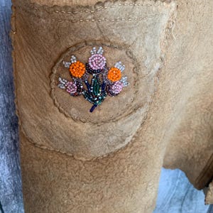 Puede incluir: Primer plano de una prenda de color beige con un bolsillo. El bolsillo presenta un parche circular con un dise&ntilde;o floral de cuentas en naranja, rosa y azul. La prenda tiene costuras visibles y una superficie texturizada.