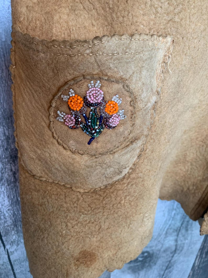 Puede incluir: Primer plano de una prenda de color beige con un bolsillo. El bolsillo presenta un parche circular con un dise&ntilde;o floral de cuentas en naranja, rosa y azul. La prenda tiene costuras visibles y una superficie texturizada.