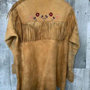 Puede incluir: Chaqueta de ante color canela con flecos en la espalda y las mangas. La espalda presenta dise&ntilde;os florales bordados en rojo, naranja y azul. La chaqueta est&aacute; colgada en una percha de madera.
