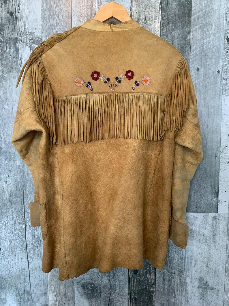 Puede incluir: Chaqueta de ante color canela con flecos en la espalda y las mangas. La espalda presenta dise&ntilde;os florales bordados en rojo, naranja y azul. La chaqueta est&aacute; colgada en una percha de madera.
