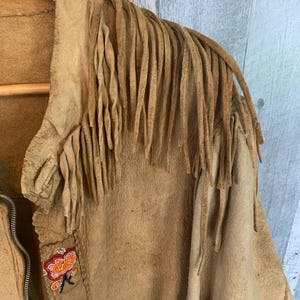 Puede incluir: Chaqueta de ante color canela con largos flecos en los hombros. La chaqueta tiene cierre de cremallera y est&aacute; decorada con abalorios florales de colores a lo largo del cuello y la parte delantera. La chaqueta est&aacute; colgada en una percha de madera.