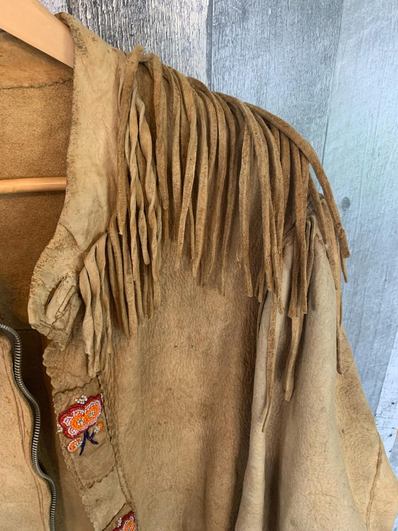 Puede incluir: Chaqueta de ante color canela con largos flecos en los hombros. La chaqueta tiene cierre de cremallera y est&aacute; decorada con abalorios florales de colores a lo largo del cuello y la parte delantera. La chaqueta est&aacute; colgada en una percha de madera.