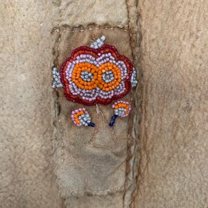 Puede incluir: Primer plano de una prenda de ante o cuero color canela con un dise&ntilde;o floral de cuentas. El dise&ntilde;o central es una flor grande, naranja y roja con peque&ntilde;os detalles de cuentas. Dos dise&ntilde;os de cuentas m&aacute;s peque&ntilde;os est&aacute;n debajo.