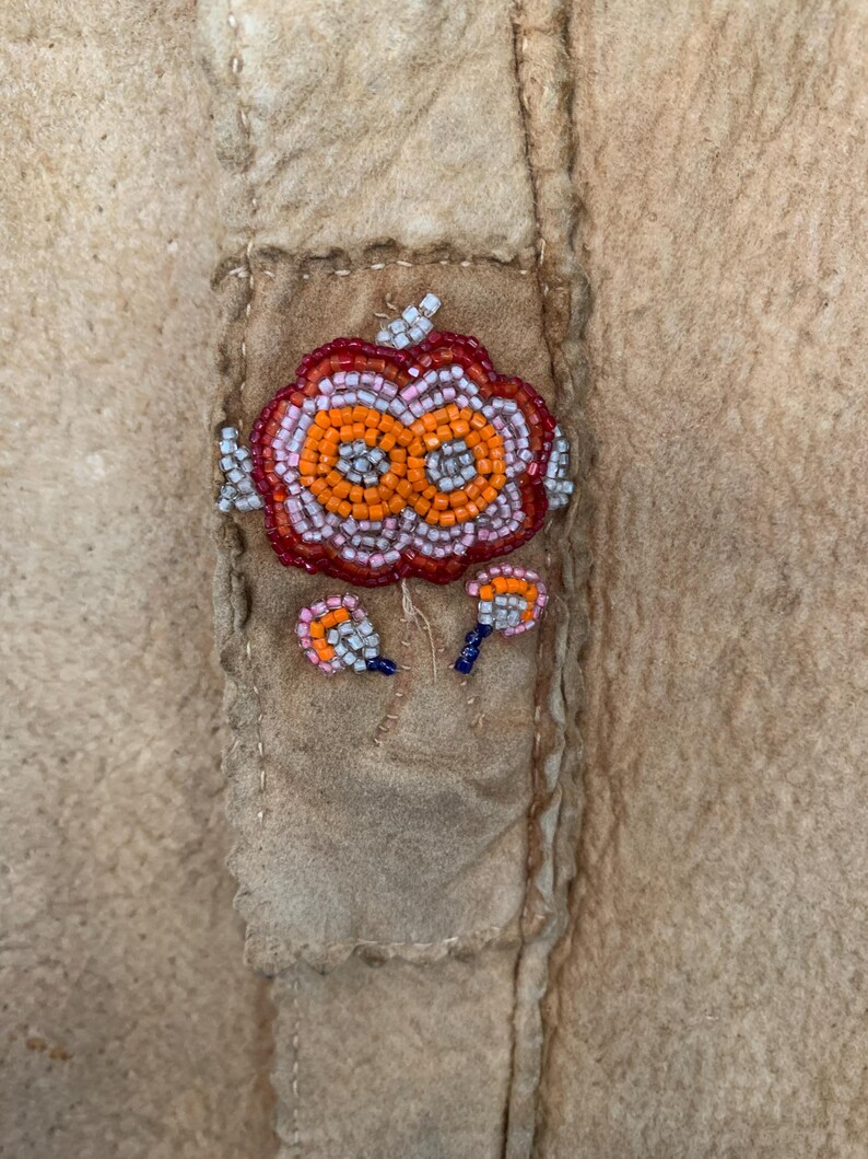 Puede incluir: Primer plano de una prenda de ante o cuero color canela con un dise&ntilde;o floral de cuentas. El dise&ntilde;o central es una flor grande, naranja y roja con peque&ntilde;os detalles de cuentas. Dos dise&ntilde;os de cuentas m&aacute;s peque&ntilde;os est&aacute;n debajo.