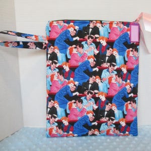 May include: A rectangular fabric pouch with a black and white patterned handle. The pouch features a repeating pattern of couples in various poses, with a pink tag and ribbon attached. The dominant colors are blue, pink, and black.