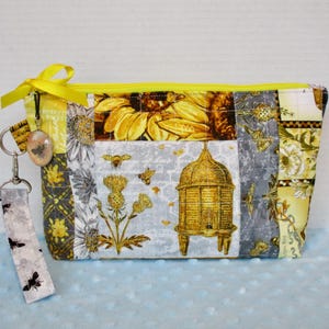 May include: A rectangular, quilted fabric pouch with a yellow zipper and a yellow bow. The pouch features a bee and honeycomb design with gray, yellow, and white floral patterns. A small charm and a wrist strap with bee graphics are attached.