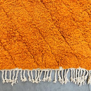 May include: Close-up of a vibrant orange rug with a textured, shaggy surface. The rug features a diamond pattern and a cream-colored fringe along one edge. The rug's warm color and texture suggest a cozy, inviting feel.