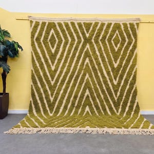 May include: A large rectangular area rug featuring a high-pile texture in a vibrant olive green colour. The rug displays a repeating geometric diamond pattern in a cream white hue, finished with long, knotted fringe along the bottom edge.