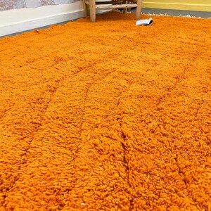 May include: A large, shaggy orange rug fills the frame, its texture appearing soft and plush. The rug's color is a vibrant, warm orange, and the fibers create a dense, textured surface. The rug is on a floor with a white baseboard and a wooden chair.