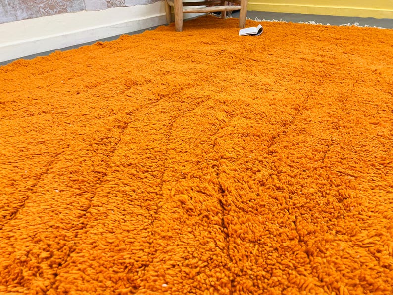 May include: A large, shaggy orange rug fills the frame, its texture appearing soft and plush. The rug's color is a vibrant, warm orange, and the fibers create a dense, textured surface. The rug is on a floor with a white baseboard and a wooden chair.