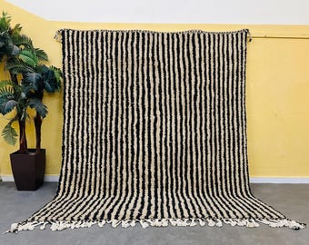 May include: A rectangular area rug featuring a repeating pattern of vertical black and cream-colored stripes. The rug has a thick, textured pile and is finished with a row of knotted cream-colored fringe along the bottom edge of the fabric.