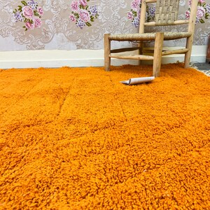 May include: An orange shag rug fills the foreground, with a woven wooden chair and a small chest in the background. A rolled-up paper rests on the rug. The wall is decorated with a floral wallpaper.