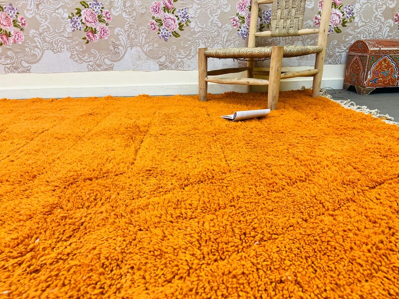 May include: An orange shag rug fills the foreground, with a woven wooden chair and a small chest in the background. A rolled-up paper rests on the rug. The wall is decorated with a floral wallpaper.
