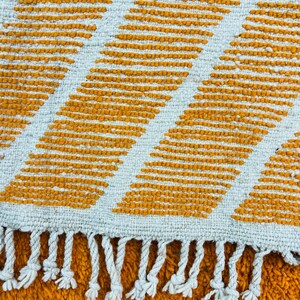 May include: Close-up of a woven rug with a geometric pattern. The rug features alternating diagonal stripes of mustard yellow and off-white. The edge of the rug has white fringe.