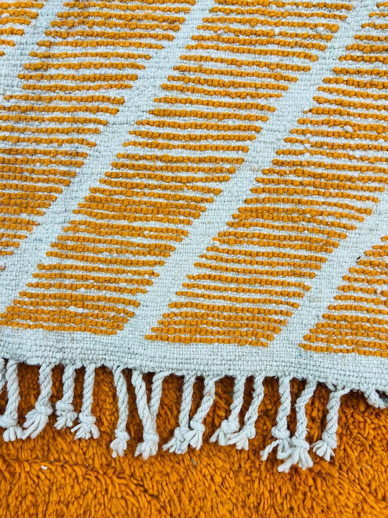 May include: Close-up of a woven rug with a geometric pattern. The rug features alternating diagonal stripes of mustard yellow and off-white. The edge of the rug has white fringe.