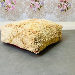 May include: A large, square, cream-colored pouf with a red trim. The pouf is made of a fluffy, textured fabric and is sitting on a gray floor.