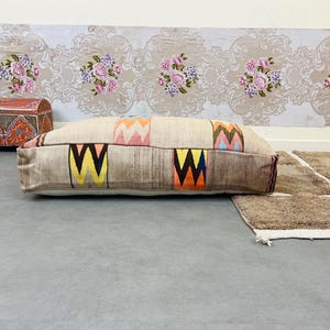 Può includere: Un grande cuscino da pavimento rettangolare e con motivi con sfondo beige e disegni geometrici colorati. Il cuscino è realizzato in tessuto tessuto e ha una texture trapuntata.