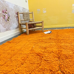 May include: An orange shag rug fills the foreground, with a woven chair and open book in the background. The walls are painted yellow and cream, with floral wallpaper and decorative accents. The rug adds a vibrant pop of color to the room.