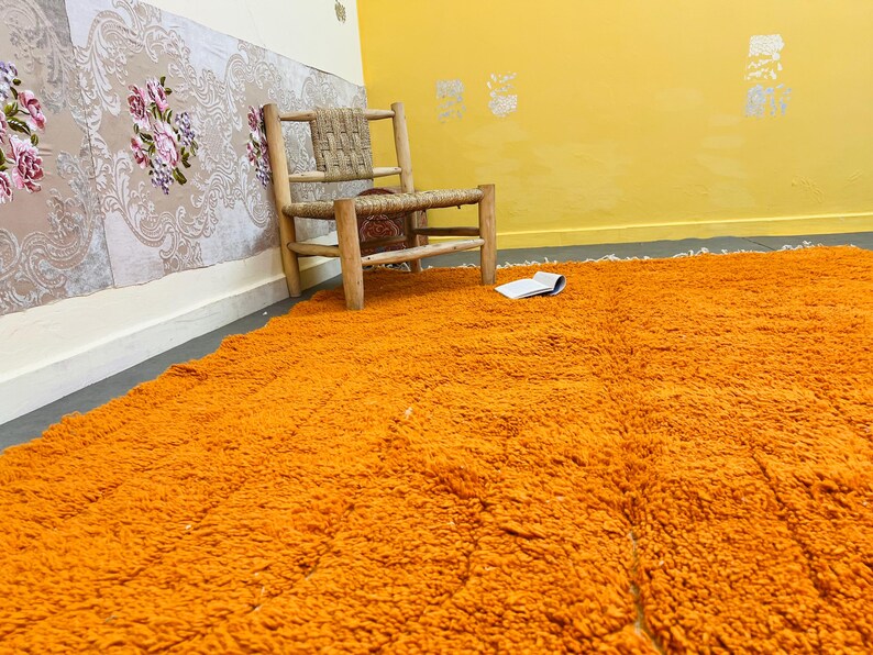 May include: An orange shag rug fills the foreground, with a woven chair and open book in the background. The walls are painted yellow and cream, with floral wallpaper and decorative accents. The rug adds a vibrant pop of color to the room.