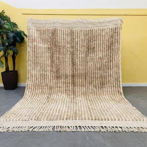May include: A rectangular area rug featuring a vertical striped pattern in alternating shades of cream and light brown. The rug has a thick, textured pile and is finished with a knotted fringe along the bottom edge, creating a soft appearance.
