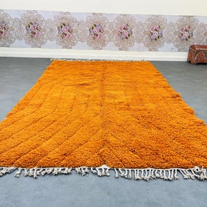 May include: A large, rectangular orange rug with a textured, shaggy surface. The rug features a geometric pattern and white fringe along the edges. The rug is placed on a gray floor.
