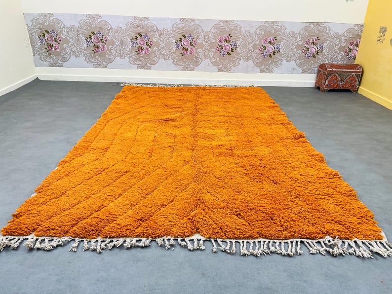 May include: A large, rectangular orange rug with a textured, shaggy surface. The rug features a geometric pattern and white fringe along the edges. The rug is placed on a gray floor.