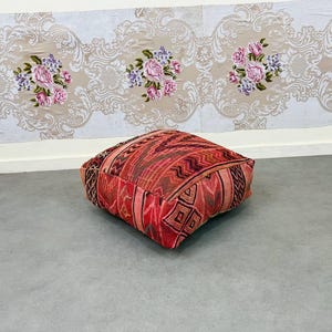 May include: A square, floor cushion with a geometric pattern in shades of red, brown, and black. The cushion is placed on a gray surface, with a floral wallpaper in the background.