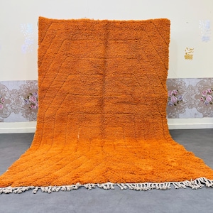 May include: A large, orange shag rug with a geometric design. The rug has a thick, textured pile and a fringe along the bottom edge. The rug is displayed against a neutral wall.