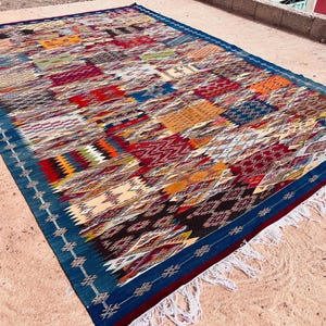 May include: A rectangular, multi-colored rug with a dark blue border and white fringe. The rug features a patchwork design with various geometric patterns in shades of red, orange, blue, and green. The rug is placed on a light brown surface.