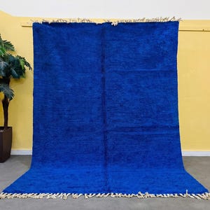 May include: A vibrant blue rug with a textured surface and a fringe along the bottom edge. The rug is displayed against a yellow wall, with a potted plant in the background. The rug's color is a deep, saturated blue.