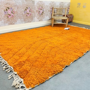May include: A large, rectangular orange rug with a shaggy texture. The rug has a cream-colored fringe along the edges. A wooden chair and a small chest are in the background, with floral wallpaper.