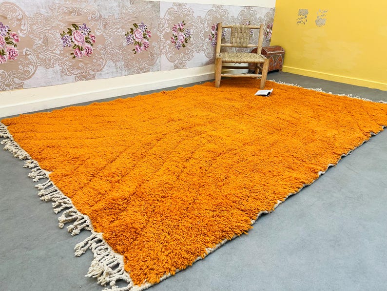 May include: A large, rectangular orange rug with a shaggy texture. The rug has a cream-colored fringe along the edges. A wooden chair and a small chest are in the background, with floral wallpaper.