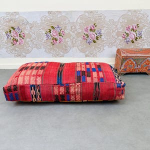 May include: A rectangular, red floor cushion with a patchwork design featuring blue, black, and pink accents. The cushion is placed on a gray floor, with a decorative chest and floral wallpaper in the background.