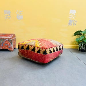 May include: A red, yellow, and black patterned floor cushion with a square shape. The cushion is made of woven fabric and has a fringed edge.