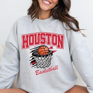 Puede incluir: Sudadera gris claro con un diseño "HOUSTON" en rojo y blanco sobre una imagen de baloncesto y red. La palabra "Basketball" está en rojo debajo. Un estilo casual.