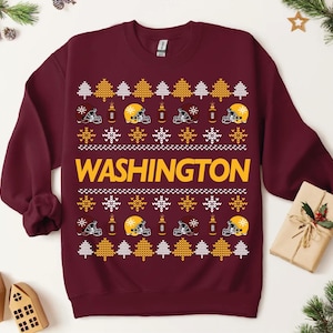 May include: Burgundy sweatshirt with a festive pattern featuring yellow football helmets, trees, and snowflakes. The word "WASHINGTON" is displayed in large yellow letters across the chest. A cozy, seasonal design.