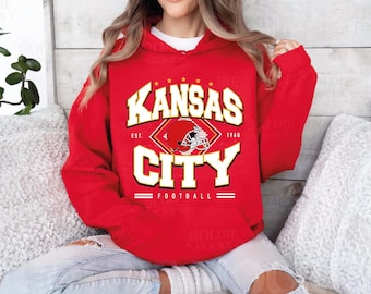 May include: Red hooded sweatshirt with a white graphic design that says "Kansas City Football" with a football helmet graphic and the text "Est. 1960".