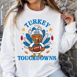 Pode incluir: Camisola branca com um desenho de peru de desenho animado usando um capacete de futebol e segurando uma bola de futebol. O texto "TURKEY AND TOUCHDOWNS" é azul. O capacete tem "DETROIT" nele. Folhas de outono e diamantes também estão no design.